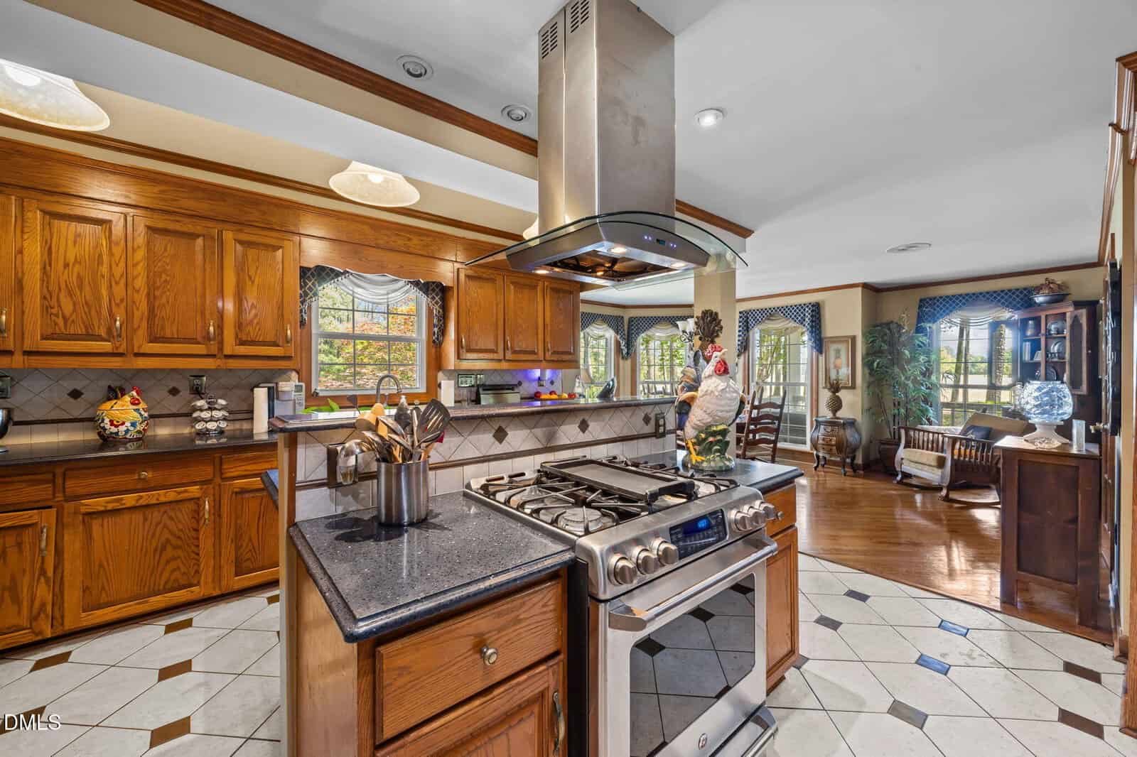 Spacious kitchen with wooden cabinets, stainless steel stove and hood, black countertops, tiled floor, and a view into a living area with large windows and various plants.