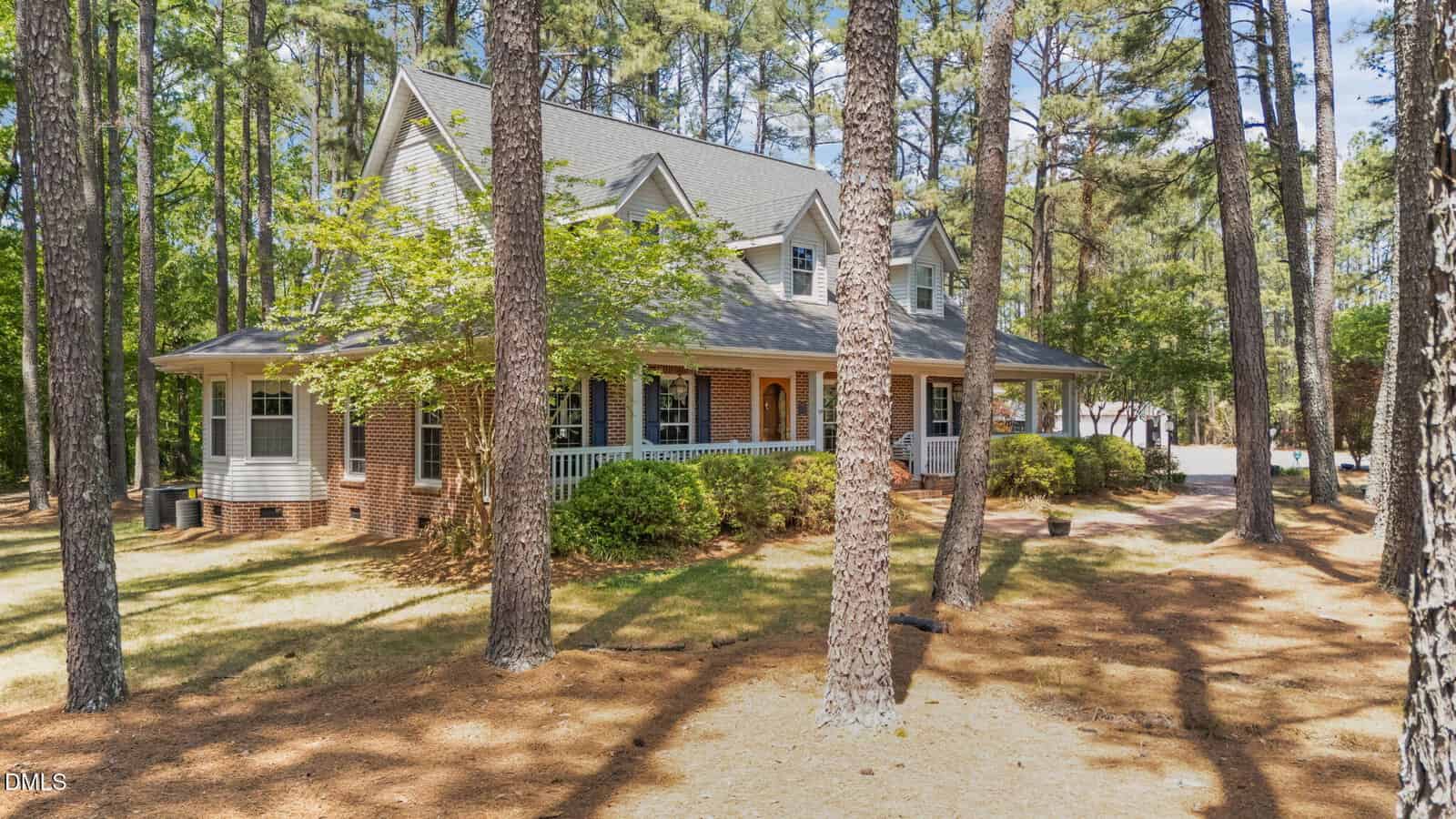 A two-story brick and siding house with a wraparound porch, surrounded by tall pine trees and set on a large, wooded lot with dappled sunlight on the ground.