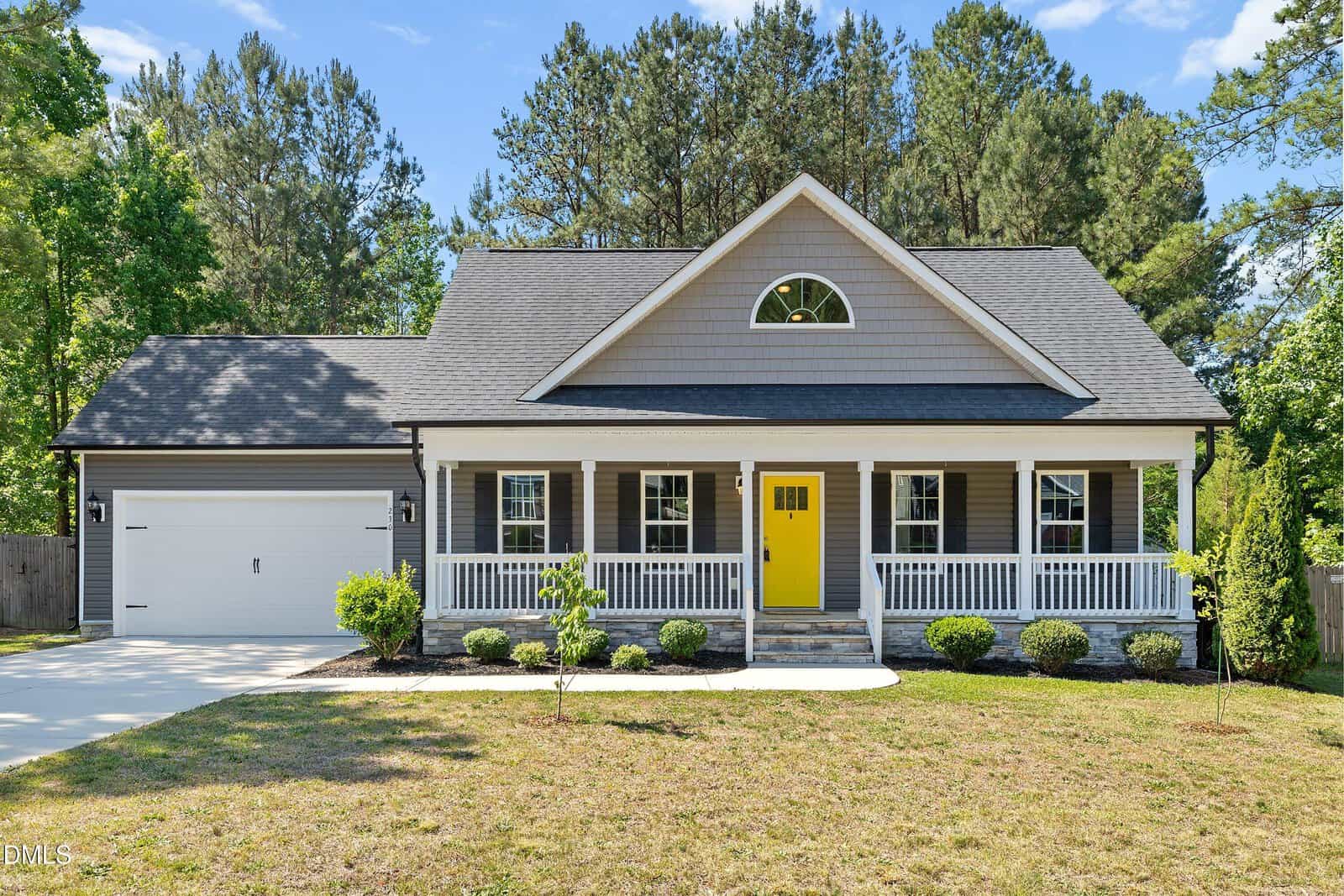A single-story house with gray siding, white trim, a yellow front door, and a covered porch. The yard is partially grassed with small shrubs, and the driveway leads to a two-car garage. Trees are visible in the background.