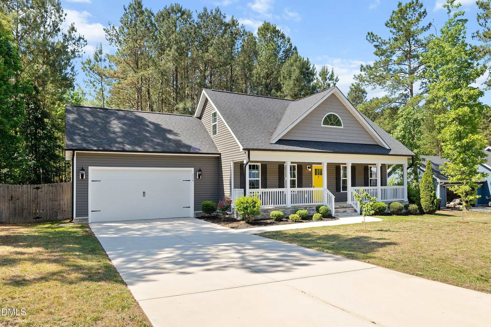 Charming single-story suburban house at 230 N Ridge Drive, Louisburg, with a front porch, attached two-car garage, manicured shrubs, and large driveway. Gray siding and white trim overlook a wooded area under a clear blue sky.