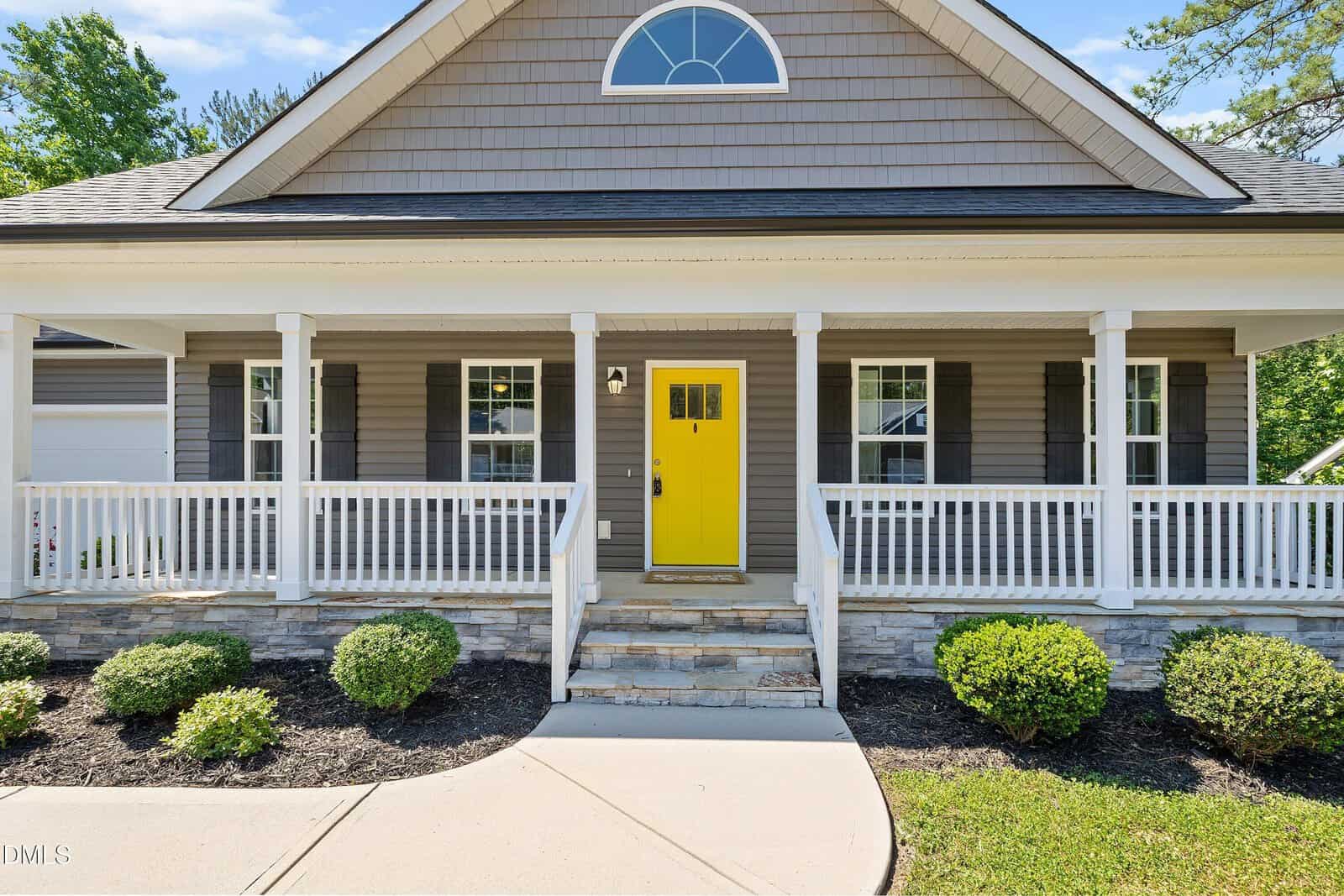 A single-story Louisburg house with gray siding, white trim, a stone foundation, a large covered front porch with white railings, and a bright yellow door at 230 N Ridge Drive. Neatly trimmed bushes line the walkway to the steps.