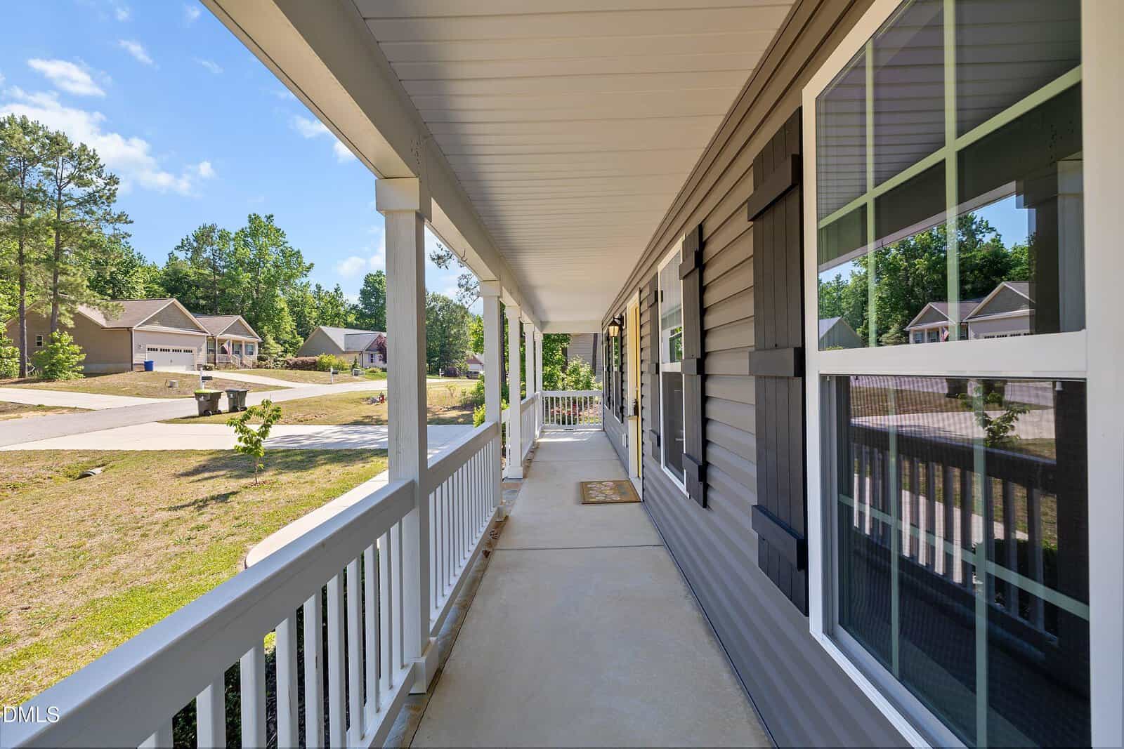 A long, covered front porch with white railings and columns, attached to a gray house with large windows, overlooks a suburban neighborhood with houses, trees, and a sunny sky.