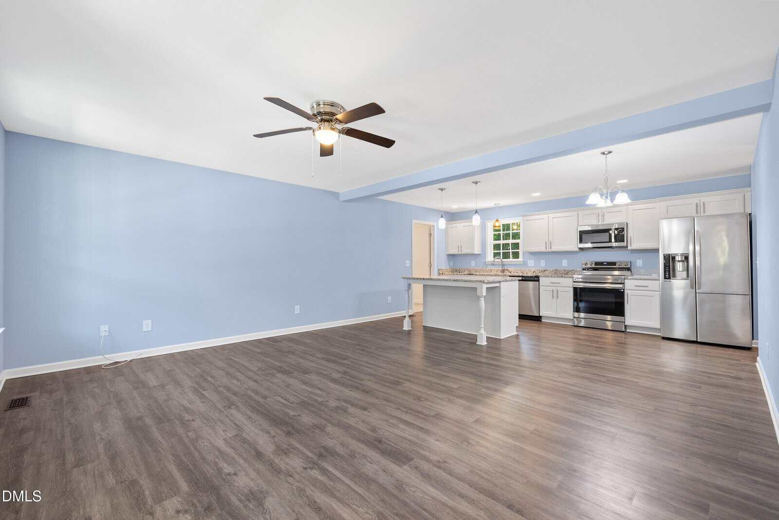 Open living area with light blue walls, dark wood flooring, and a ceiling fan. The modern kitchen features white cabinets, stainless steel appliances, and an island with pendant lighting.