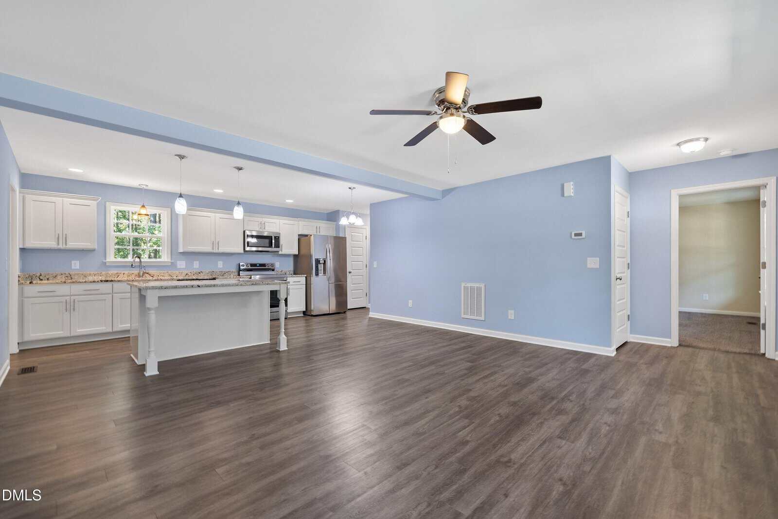 Open-concept kitchen and living area with wood flooring, light blue walls, white cabinets, stainless steel appliances, a center island, ceiling fan, and recessed lighting. A doorway leads to an adjacent carpeted room.