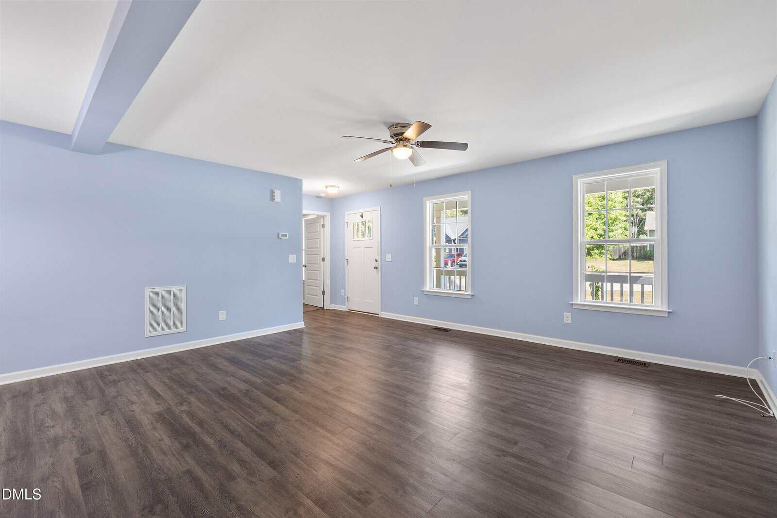 A spacious, empty living room with light blue walls, dark wood flooring, two large windows, a ceiling fan, and a view of the adjacent entryway and front door.