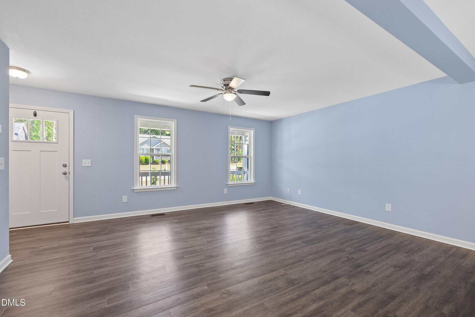 Spacious living room with light blue walls, dark wood flooring, two windows, a white front door with glass panels, and a ceiling fan with lights. Natural light brightens the empty room.