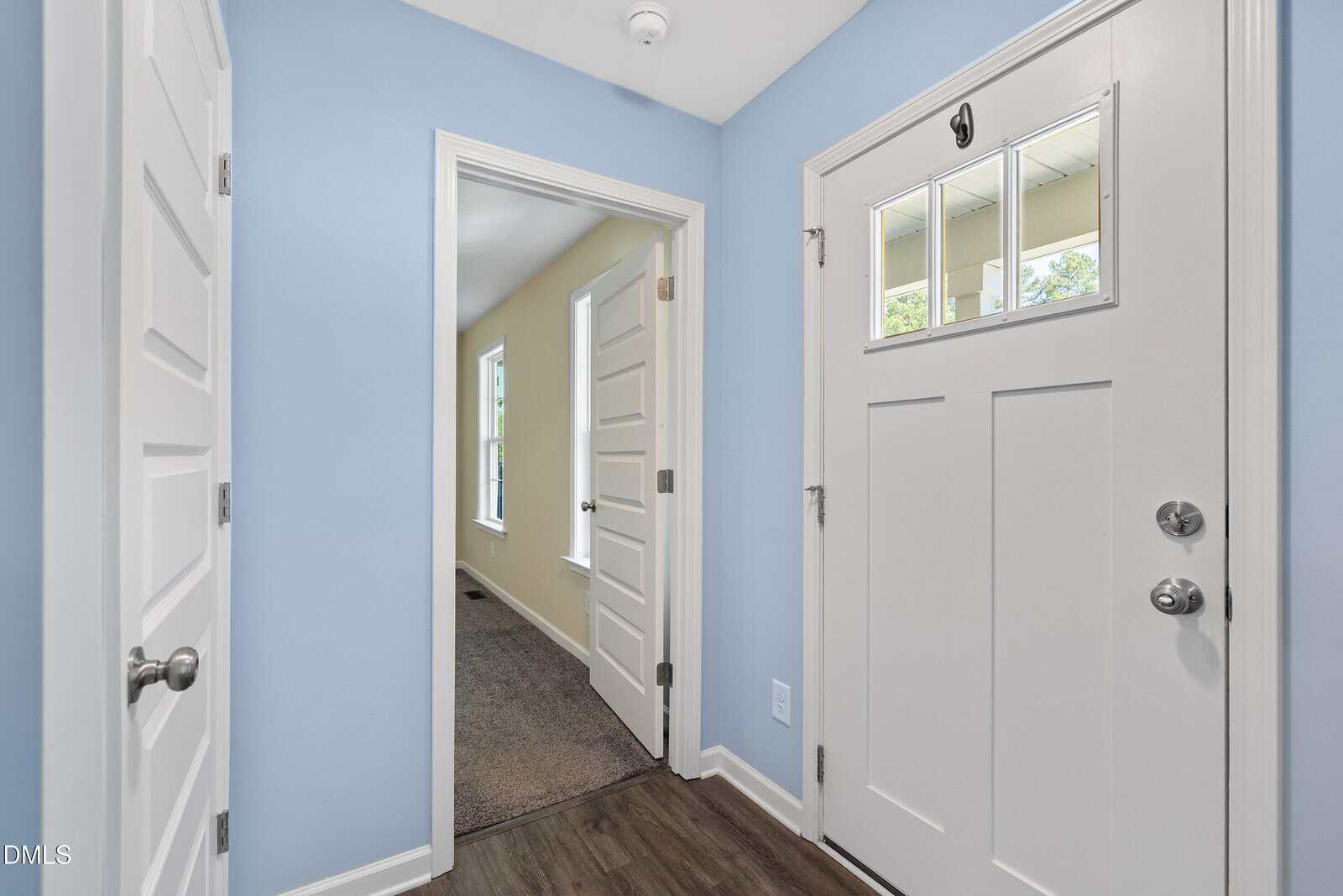 A small entryway with light blue walls, a white front door with glass panes, white interior doors, and wood flooring. An adjacent room with beige walls and carpet is visible through an open doorway.