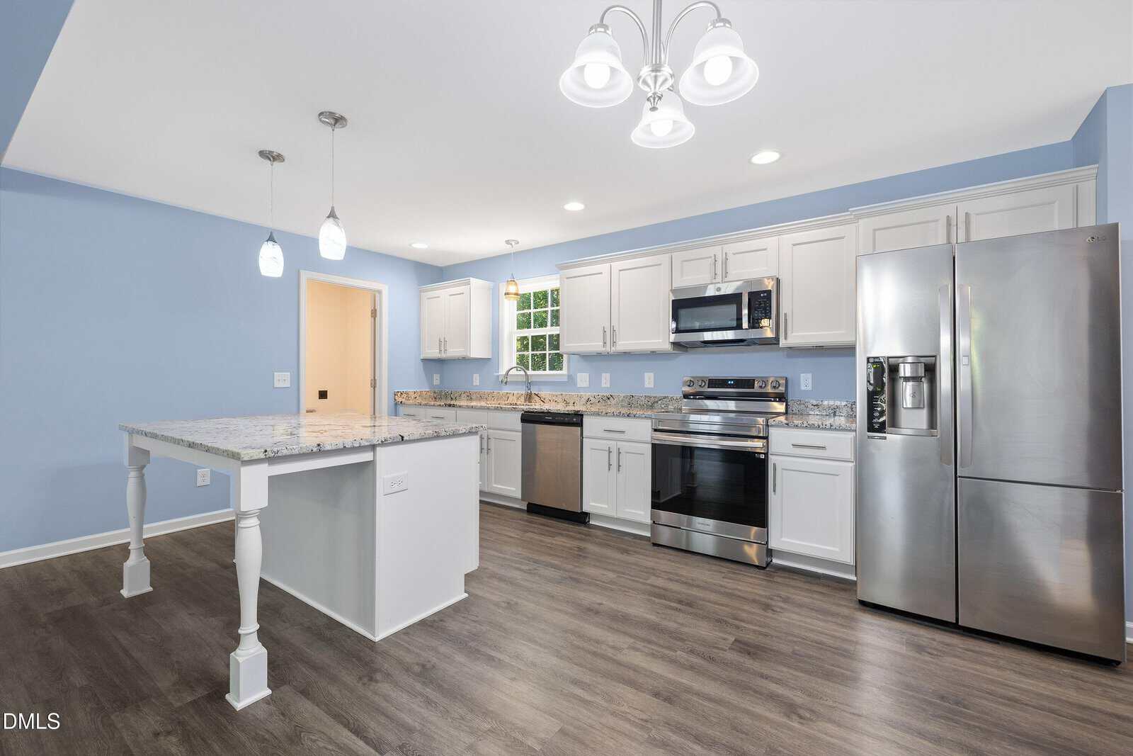 Modern kitchen with light blue walls, white cabinets, stainless steel appliances, marble island with pendant lights, and wood flooring. A window above the sink lets in natural light.