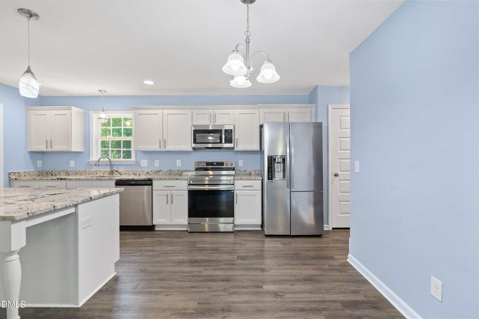 Modern kitchen with white cabinets, stainless steel appliances, granite countertops, pendant and ceiling lights, wood flooring, and light blue walls. A window above the sink lets in natural light.