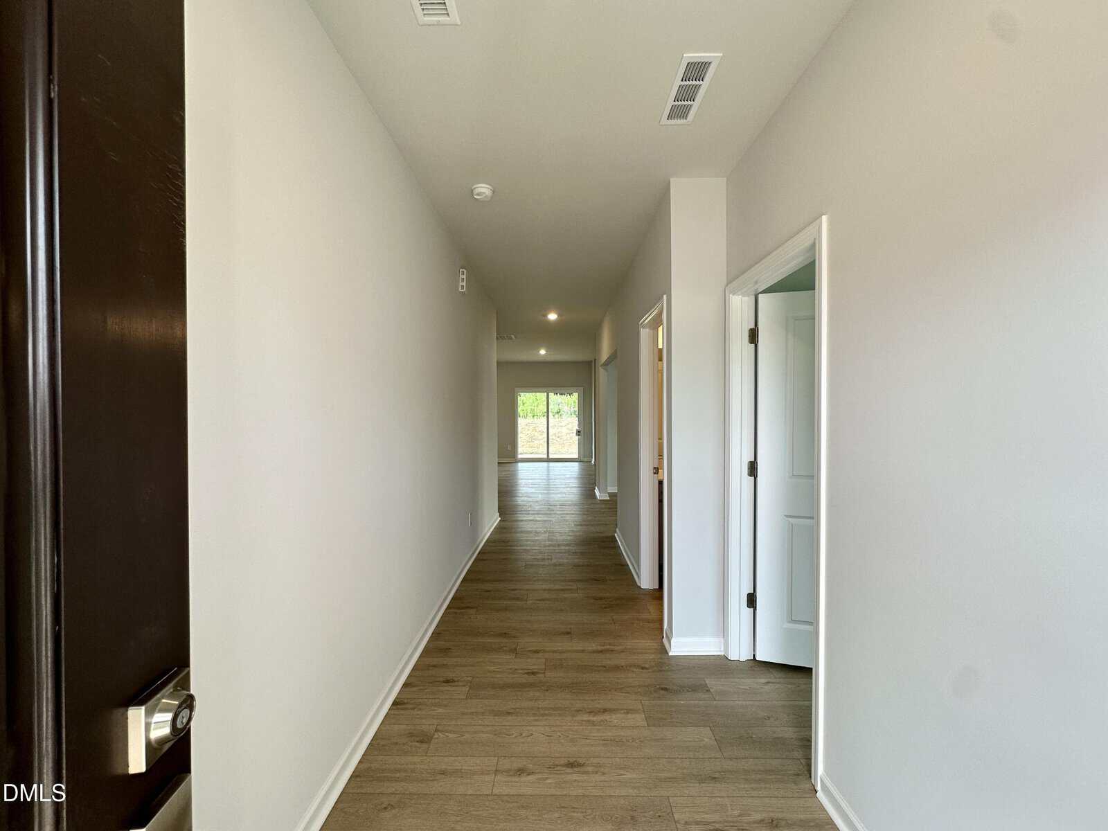 View of a long, narrow hallway with wood flooring and light-colored walls; open doorways on the right and a distant door leading outside with natural light coming in.