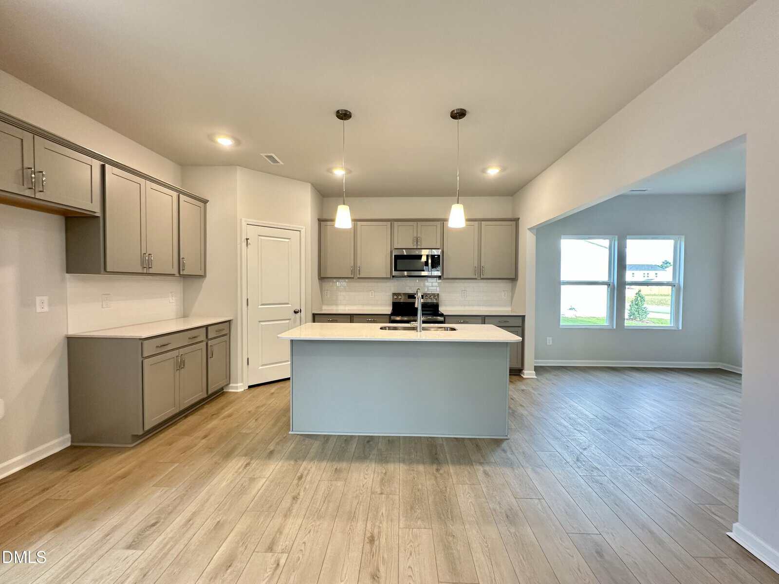 Modern kitchen with gray cabinets, a center island, stainless steel appliances, pendant lights, light wood flooring, and an adjacent dining area with large windows.