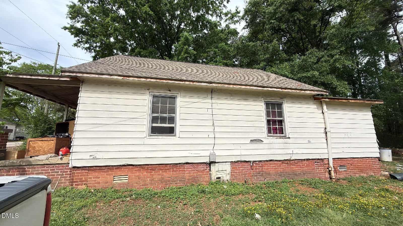 A small, single-story house with white wooden siding, a brick foundation, and a shingled roof. The house has two windows and sits in a grassy yard with some trees in the background.