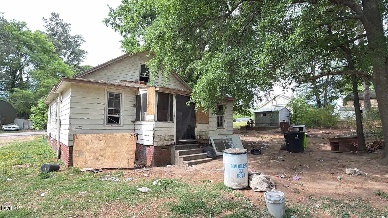 An abandoned, boarded-up house with broken windows and peeling paint sits under a large tree. The yard is littered with trash, debris, and a blue trash bin. Another small building is visible in the background.