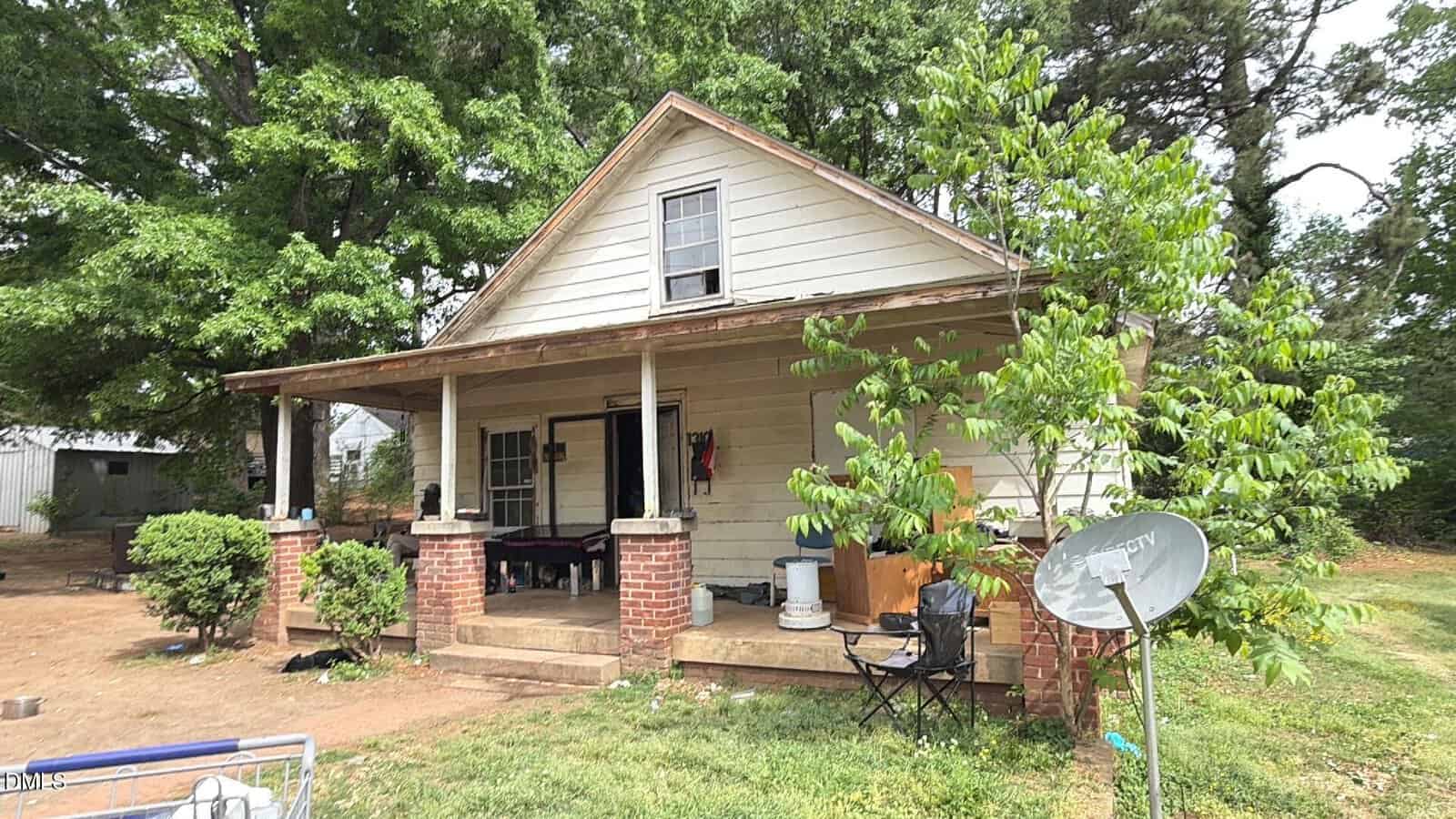 A small, single-story house at 1310 2nd Street in Henderson features a covered front porch with brick pillars and white siding. A satellite dish and chair sit in the yard, surrounded by trees and bushes. The porch holds various items and outdoor furniture.