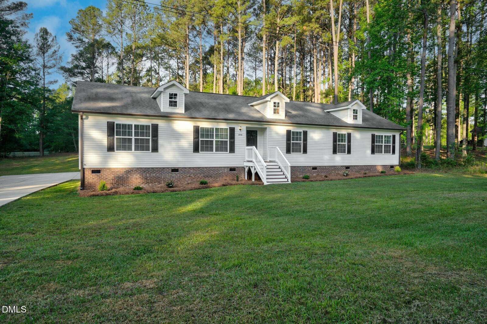 This single-story house at 2790 Sledge Road, Louisburg features white siding, a brick foundation, and three dormer windows. It sits on a spacious grassy lawn with tall trees and welcoming white steps leading up to the front door.