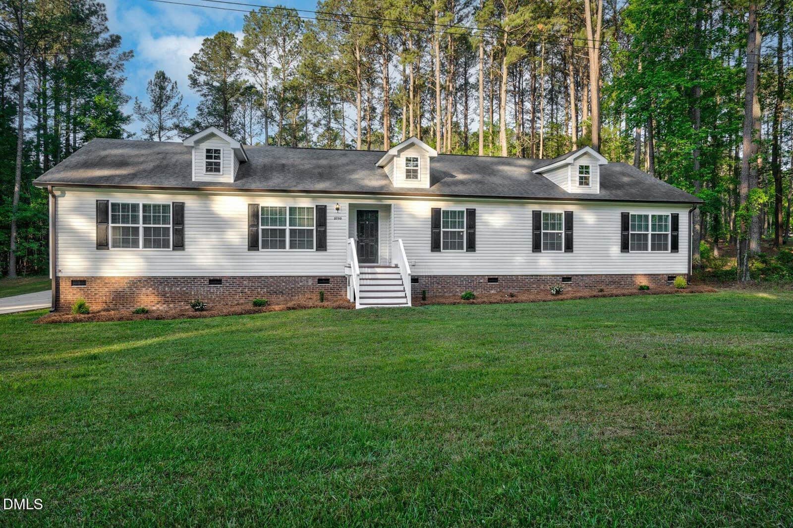 A single-story white house with a gray roof, brick foundation, and three dormer windows sits on a large, green lawn surrounded by tall trees. White steps lead to the front door at the center of the house.