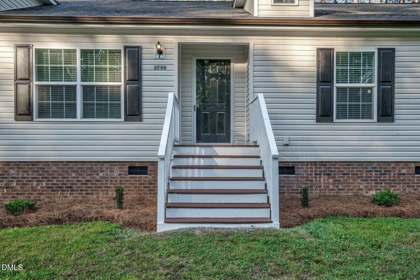 Front view of a house entrance with a glass door, white steps with railings, brick foundation, beige siding, black shutters on windows, and neatly landscaped yard.