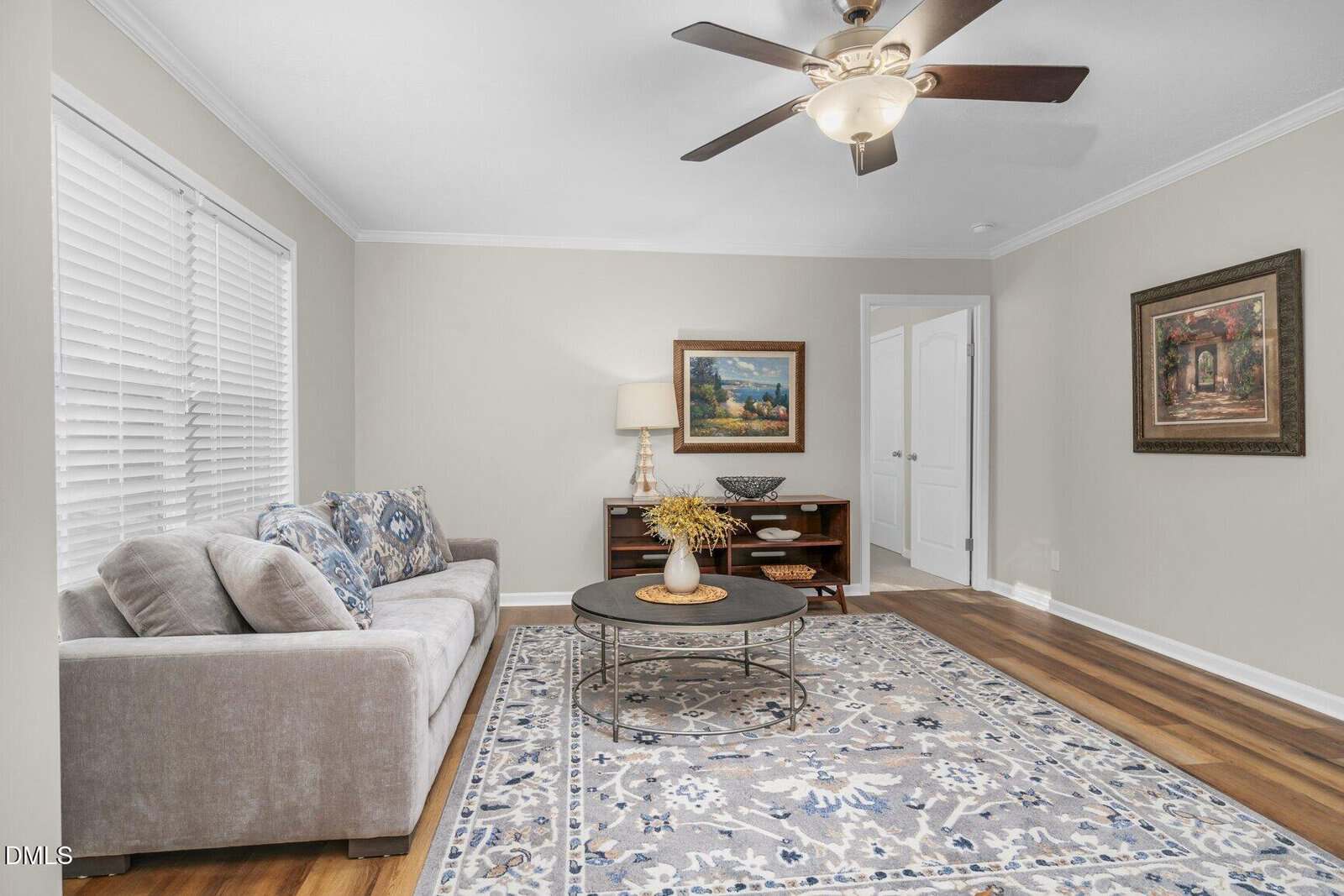 A cozy living room with a gray sofa, patterned cushions, a round coffee table on a blue and gray rug, a wooden console table with a lamp and decor, framed art on the walls, and a ceiling fan overhead.
