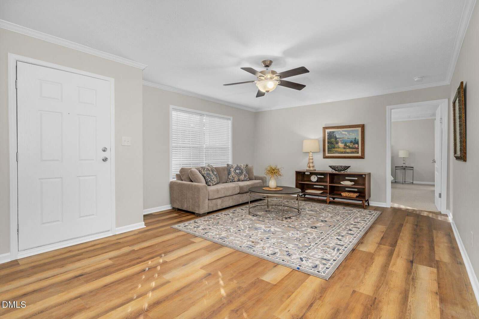 A bright living room with wood flooring, a beige sofa, patterned rug, round glass coffee table, and a wooden console under a painting. A ceiling fan with light hangs above, and sunlight streams through a window.
