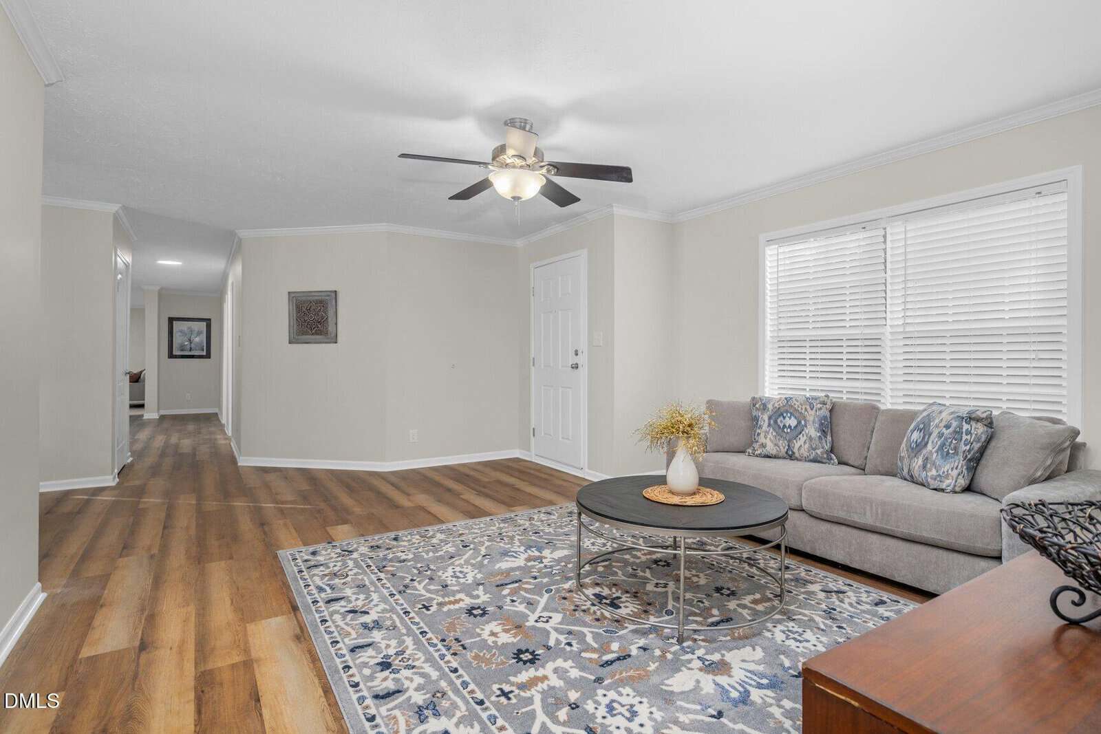 Bright living room with wood flooring, gray sectional sofa, patterned blue and white rug, coffee table, ceiling fan, and large window with blinds; hallway visible in the background.