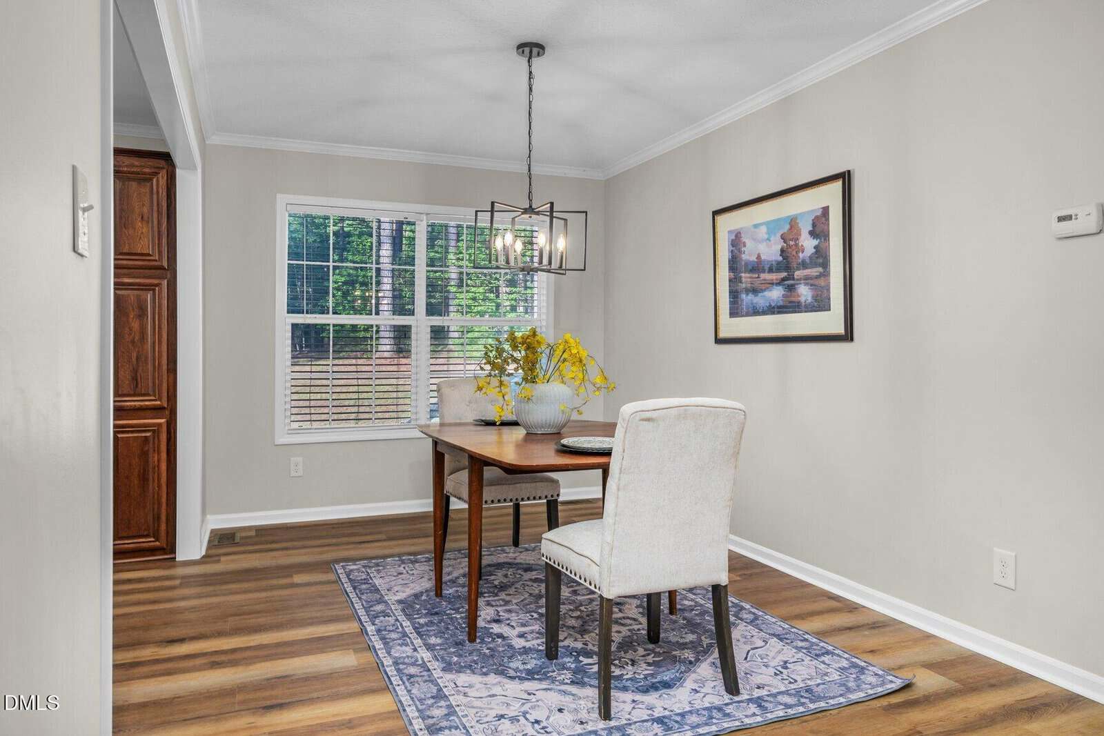 A small dining area with a wooden table, two cream chairs, yellow flowers in a vase, a blue rug, a pendant light, a window with blinds, and a framed artwork on a beige wall.