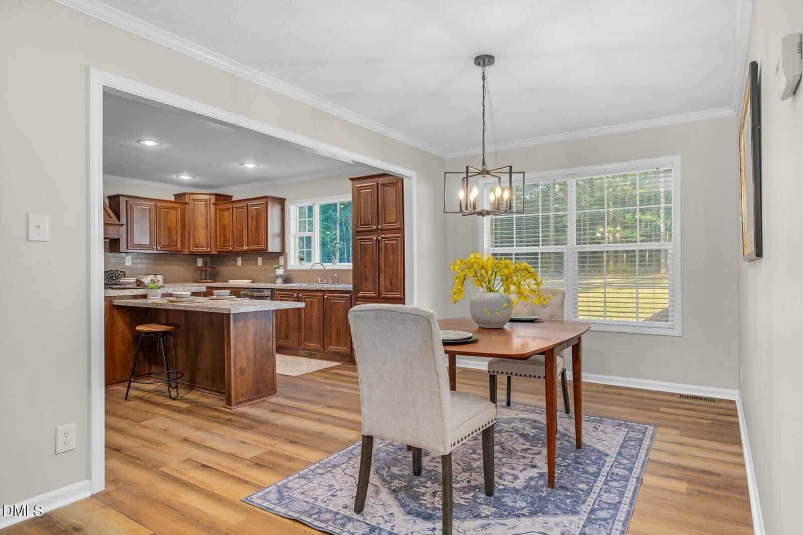 A dining room with a wooden table and two chairs sits next to a kitchen with brown cabinets and a breakfast bar. Large windows let in natural light, highlighting the wood flooring and decorative rug.