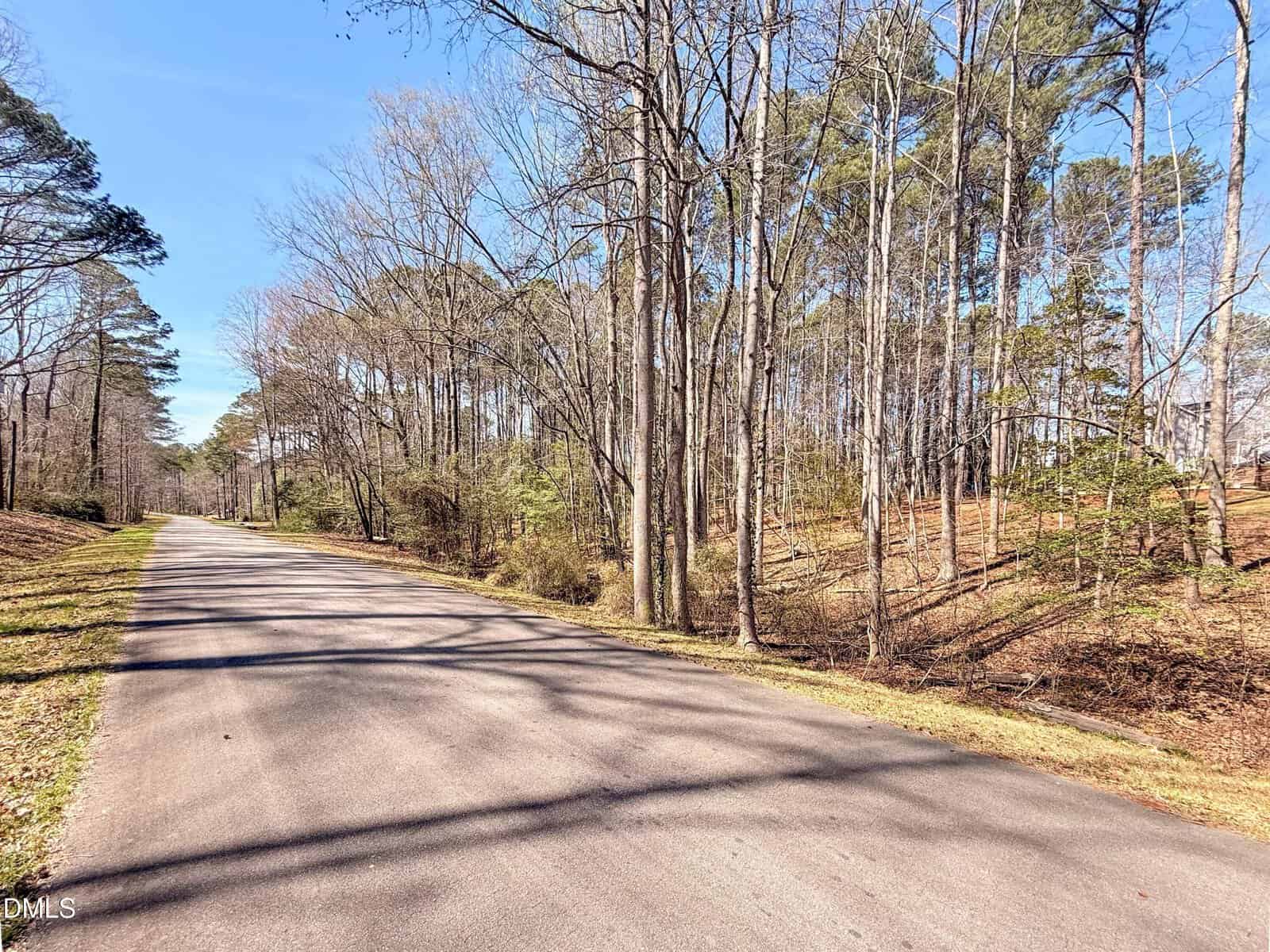 A paved road runs through a wooded area with tall, leafless trees on a sunny day at 131 Cherokee Drive, Louisburg. Shadows from the trees fall across the road, and the sky is clear and blue.