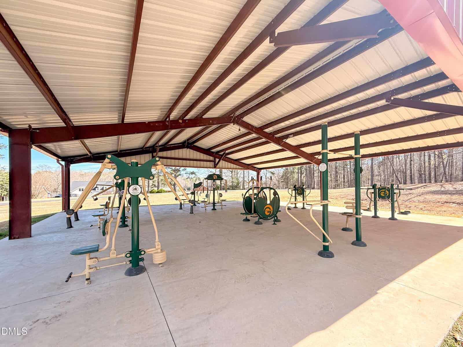 Covered outdoor gym area with various green and beige exercise equipment on a concrete slab, surrounded by grass and trees, under a metal roof with exposed beams.