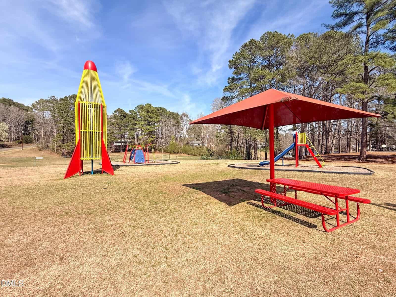A playground with a red-and-yellow rocket climbing structure, swings, a red picnic table under a red canopy, trees in the background, and a clear blue sky overhead.