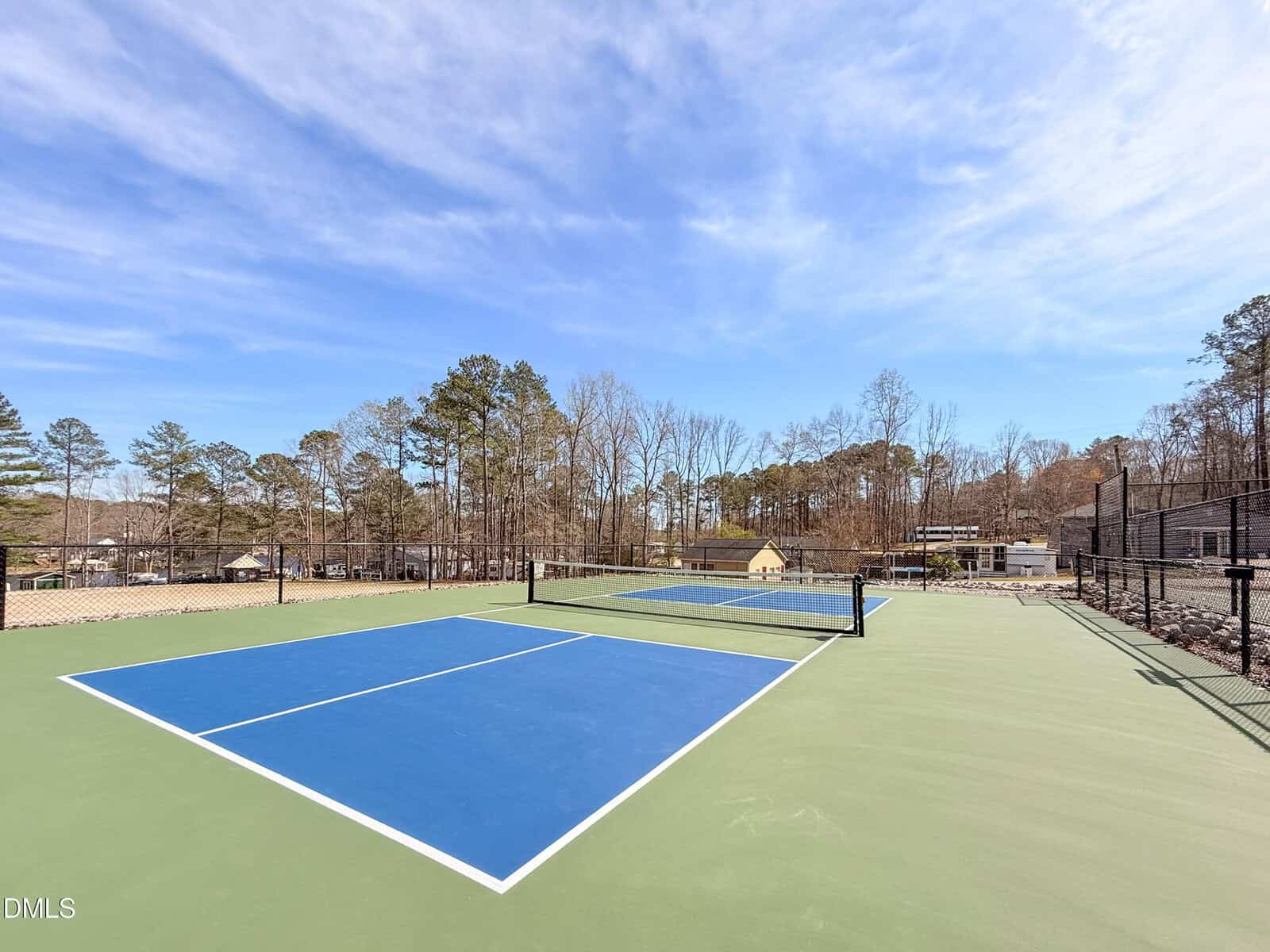 An empty outdoor tennis court with a blue playing surface and green borders, surrounded by a black chain-link fence, trees, and houses in the background under a partly cloudy sky.
