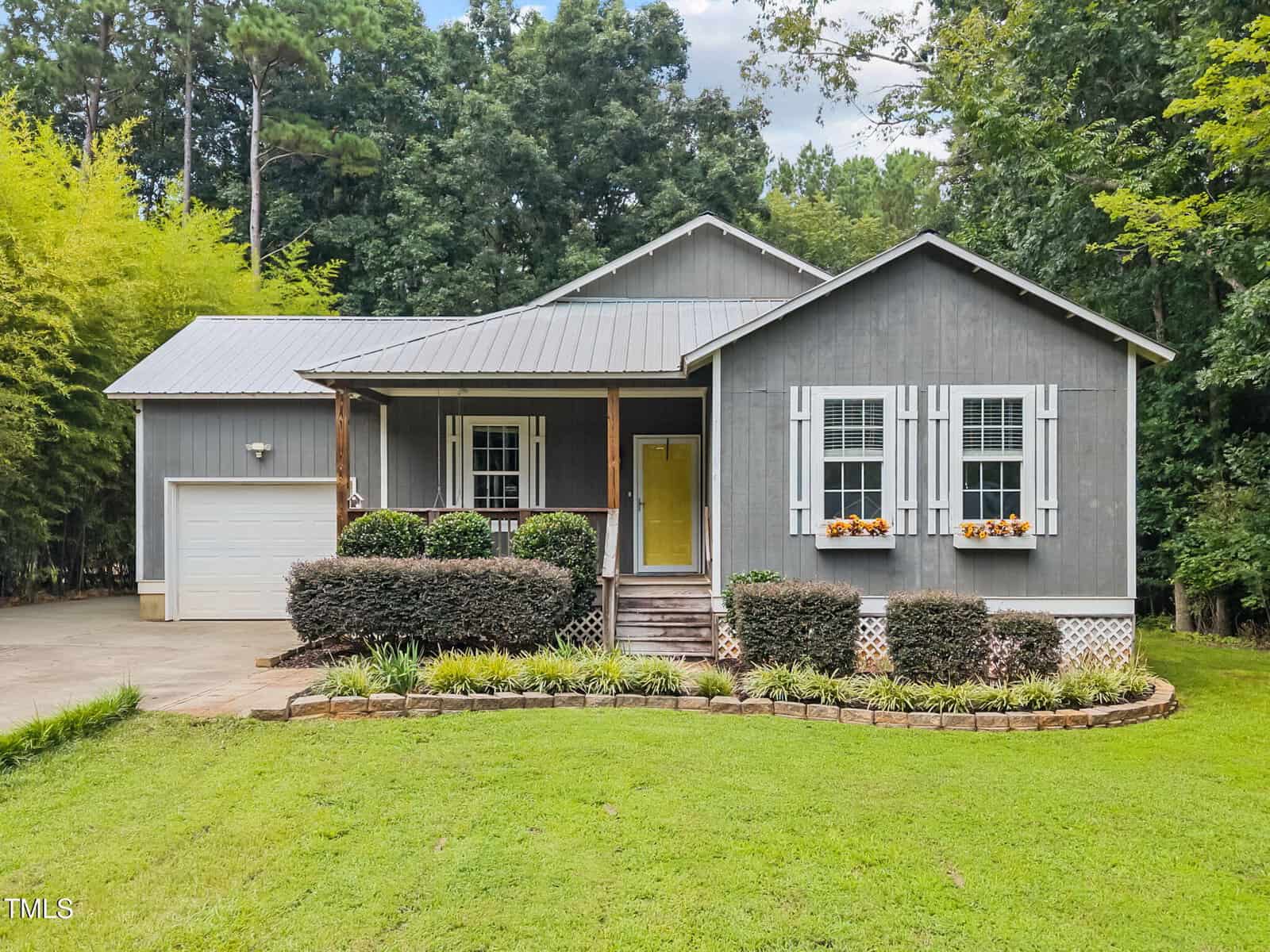 A gray single-story house with white trim at 121 Running Bear Drive, Louisburg, features a yellow front door, flower boxes under the windows, a covered porch, manicured bushes, and a two-car garage set against trees and a green lawn.