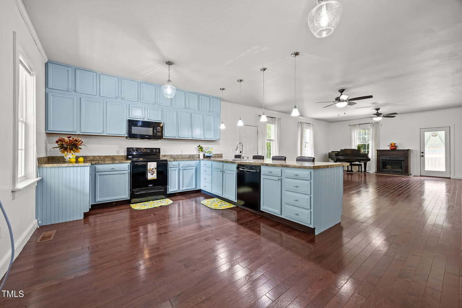 Spacious kitchen with light blue cabinets, dark appliances, and a large island, open to a living area with wood floors, ceiling fans, a piano, and a fireplace. Large windows provide natural light.