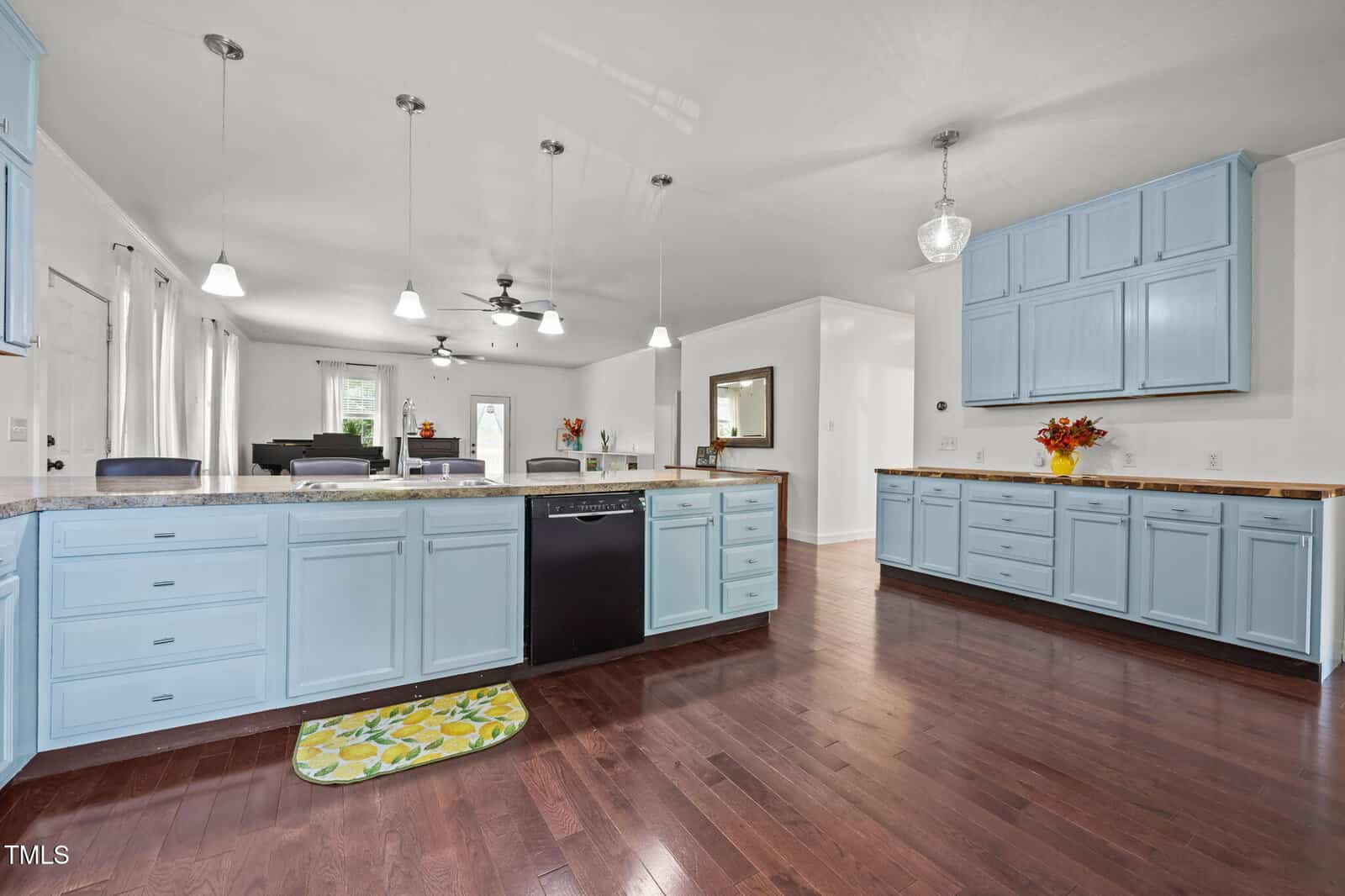 Spacious kitchen with light blue cabinets, dark appliances, wooden countertops, and dark hardwood floors. The room is brightly lit with pendant lights and natural light from large windows. Dining and living areas are visible in the background.
