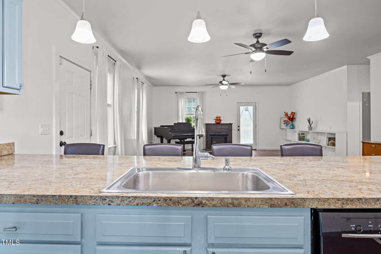 View from a kitchen island with a sink, looking into a bright living room featuring a piano, ceiling fans, large windows with curtains, and a built-in shelf with decor.