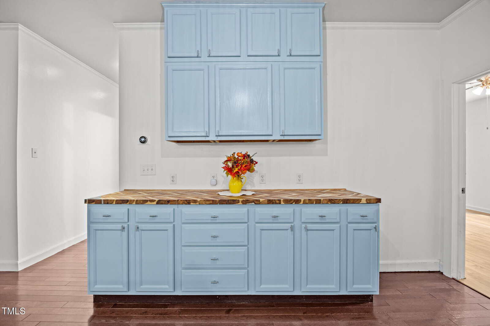 Light blue kitchen cabinets with a wood countertop, featuring a small vase of orange and red flowers. The cabinets are set against a white wall with hardwood flooring.