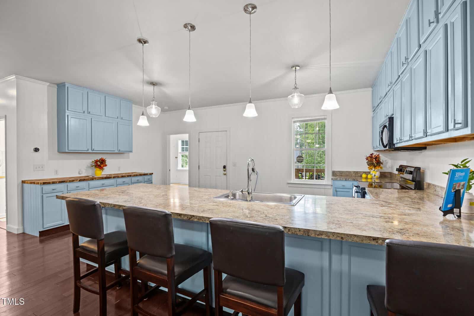 Bright kitchen with light blue cabinets, granite countertops, pendant lights, a large island with a sink, and four dark wooden barstools. Autumn decorations are placed on the counters near the walls and windows.