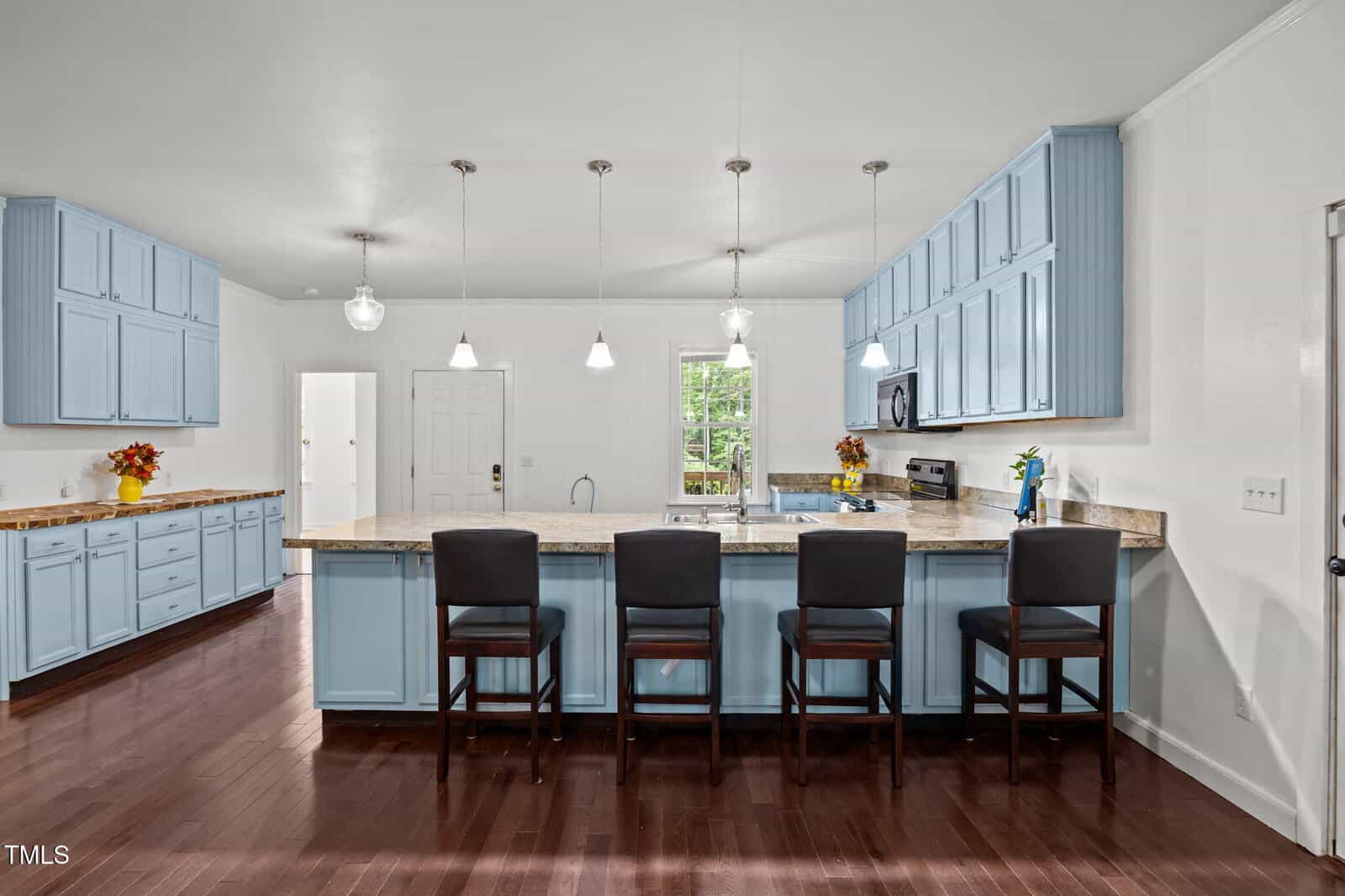 Modern kitchen with blue cabinets, granite countertops, three pendant lights, four dark barstools, hardwood floors, and vases of flowers on the counters. A window and a door are visible in the background.