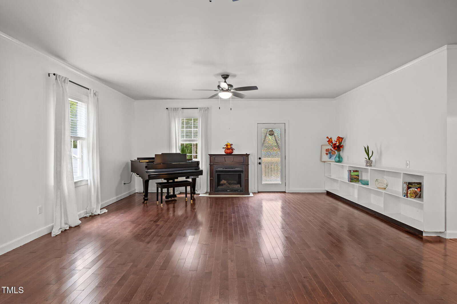Spacious living room with hardwood floors, a black grand piano, fireplace, ceiling fan, built-in shelves with decor, white curtains, and a door leading outside.