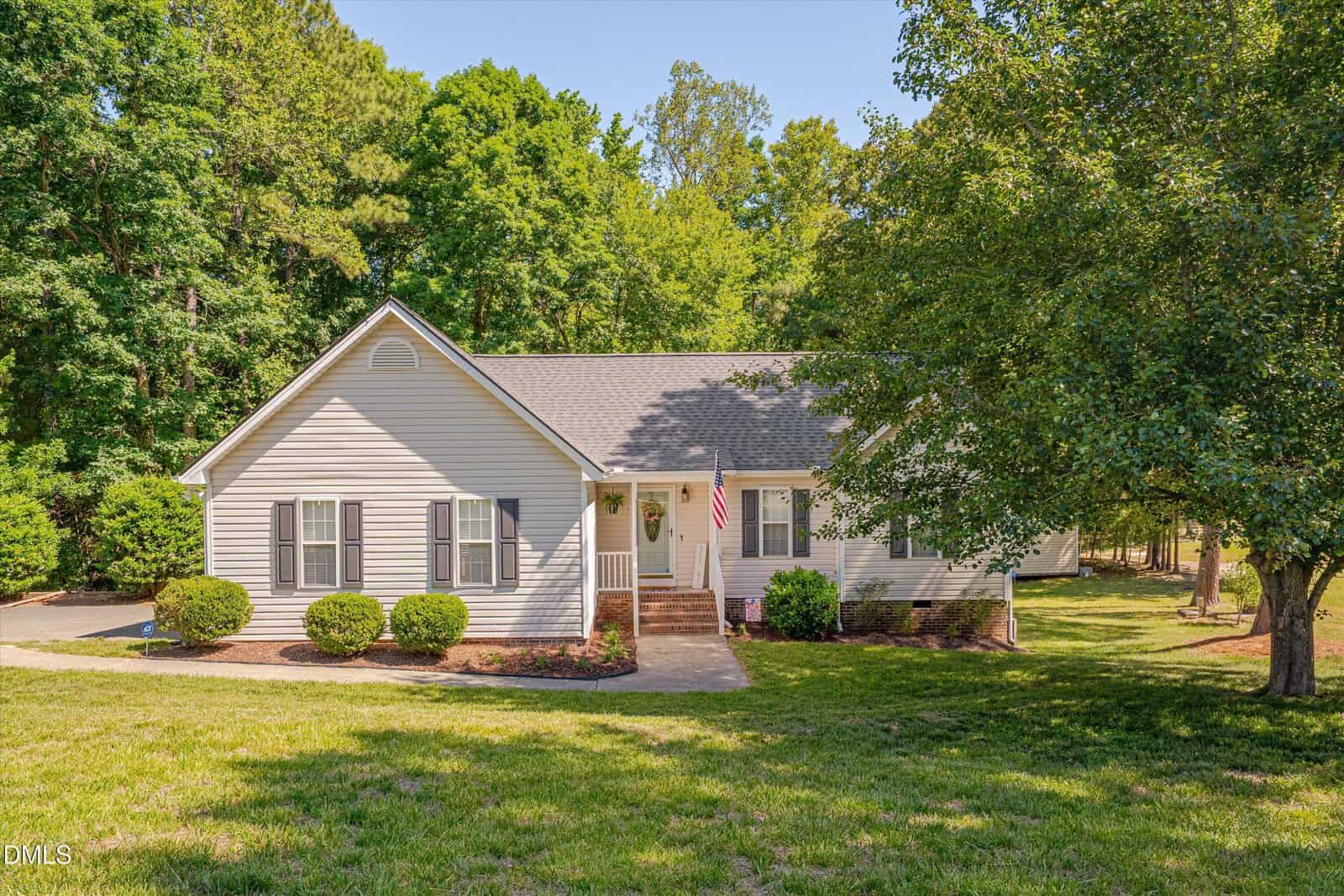 Charming single-story house at 131 N Cobble Creek Drive in Henderson with light-colored siding, a front porch, green shutters, and an American flag. The home is surrounded by a well-kept lawn, small shrubs, and large trees under a clear sky.