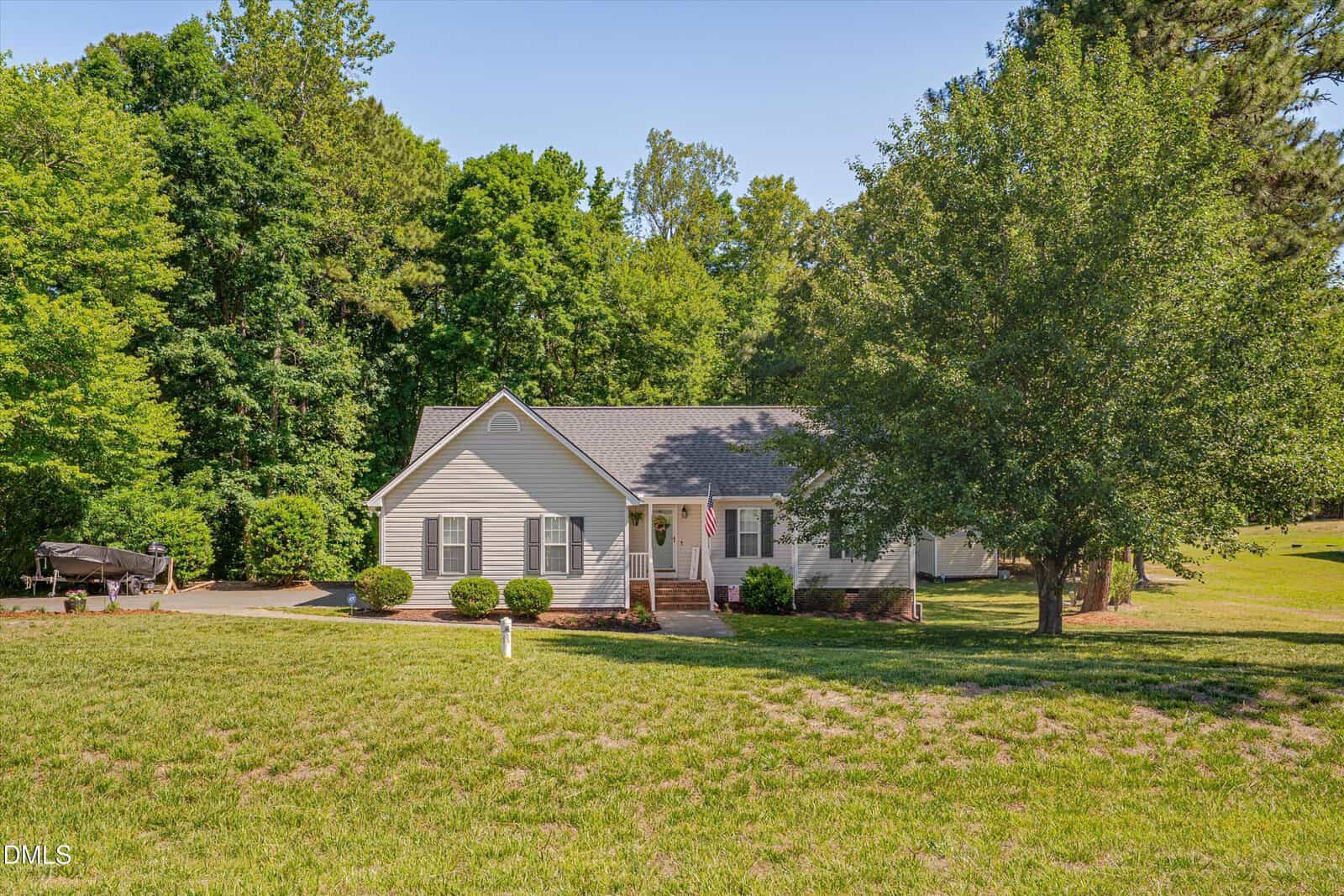 Single-story beige house with gray roof, front porch, and American flag, surrounded by green trees and a spacious grassy yard. A boat is parked on a trailer to the left of the house.