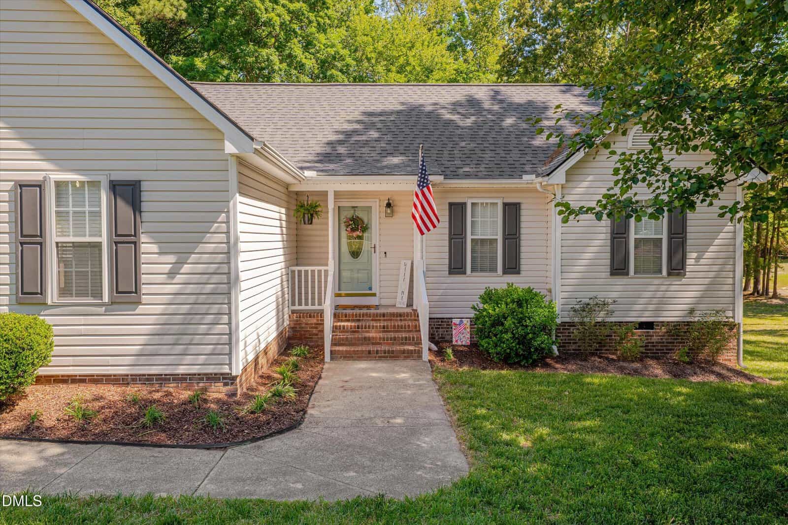 Single-story house with light gray siding, black shutters, a front porch with white railings, potted plants, an American flag, brick steps, and landscaped bushes along the walkway.