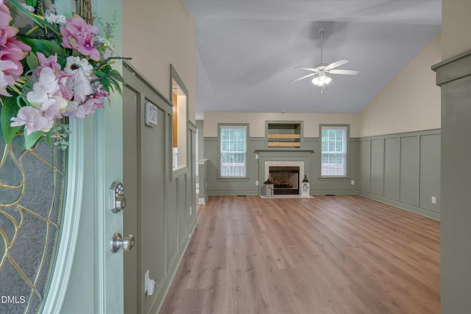 A spacious living room with green wainscoting, light wood flooring, a ceiling fan, and a fireplace. The door is partially open, decorated with pink and white flowers. Large windows let in natural light.