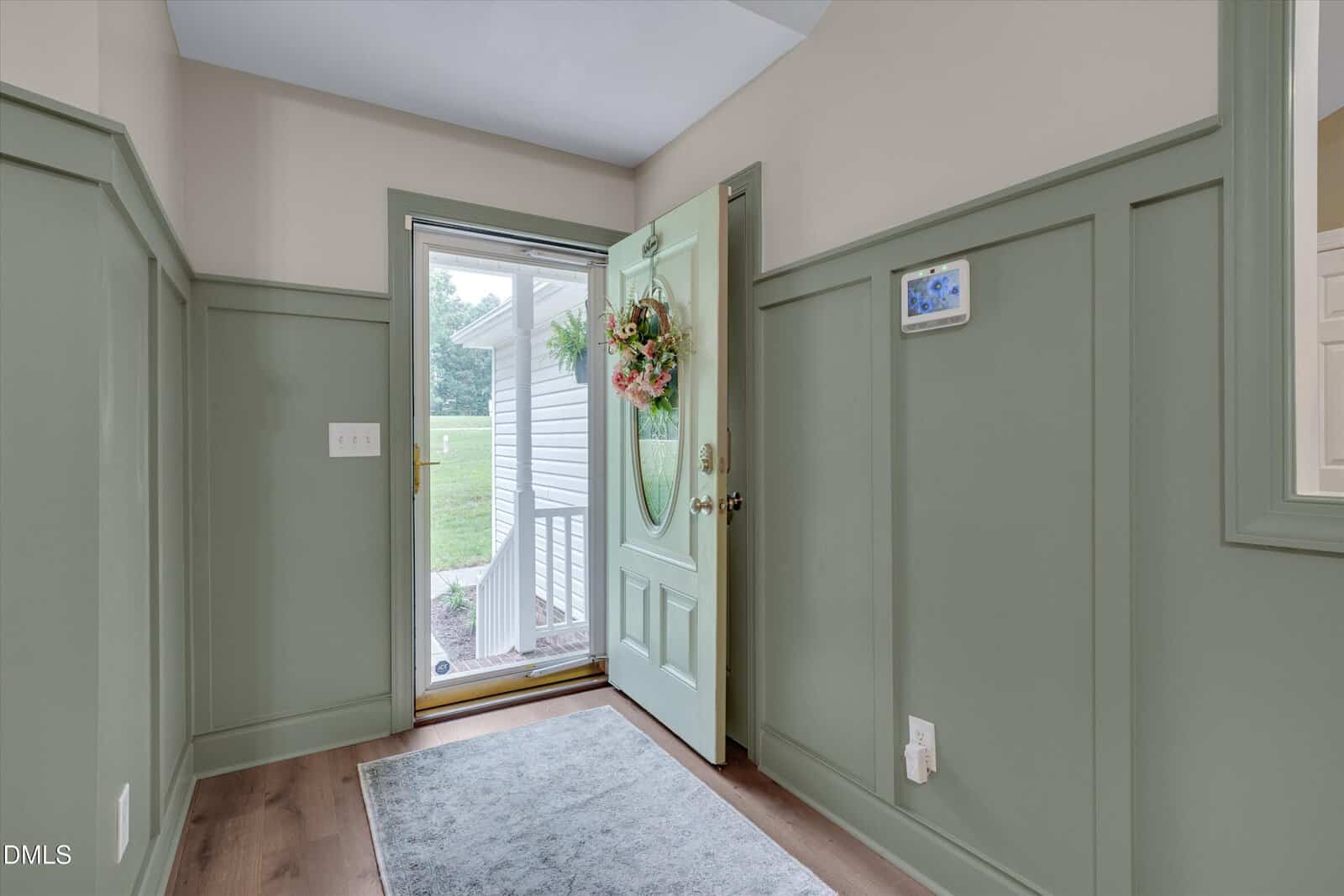 A front door painted green is open, revealing a porch with white railing and greenery outside. A floral wreath hangs on the door, and a gray rug lies on the wooden floor inside the entryway.