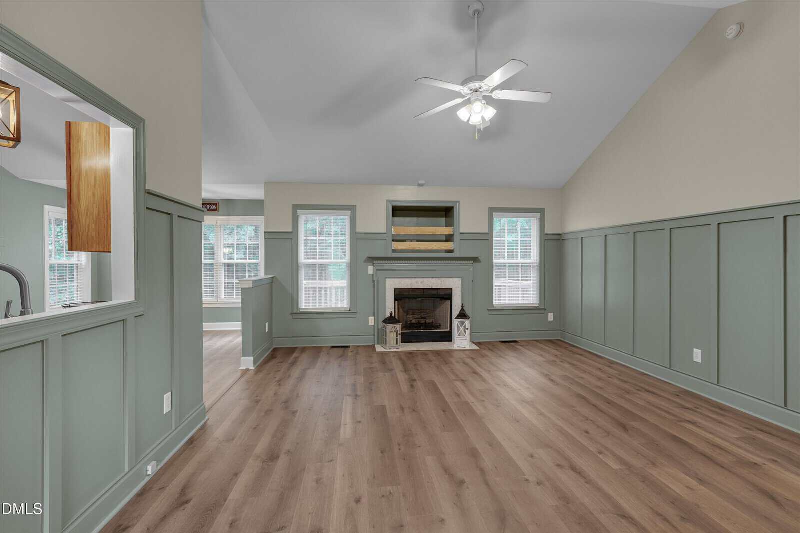 Spacious living room with light wood flooring, sage green wainscoting, a white ceiling fan, and a fireplace centered on the far wall, flanked by built-in shelves and large windows.