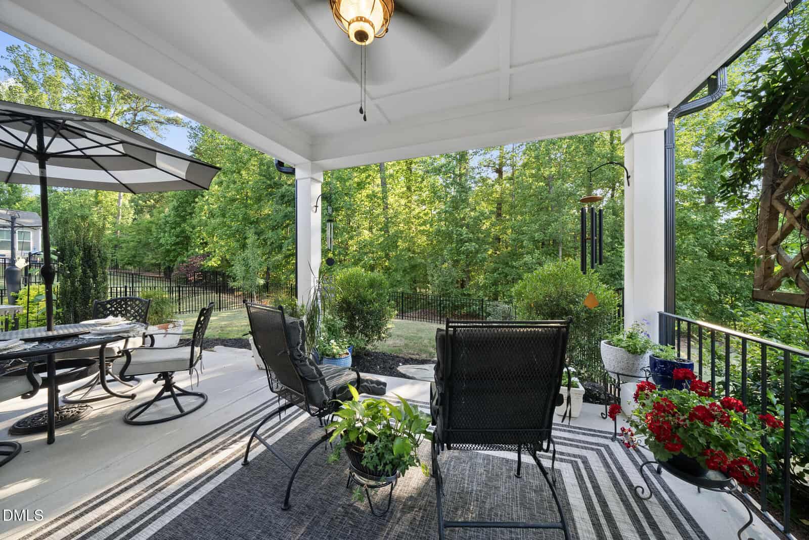 Covered patio with black chairs, a striped rug, potted plants, and a ceiling fan, overlooking a green fenced backyard with trees, an outdoor dining table, and red flowers near the railing.