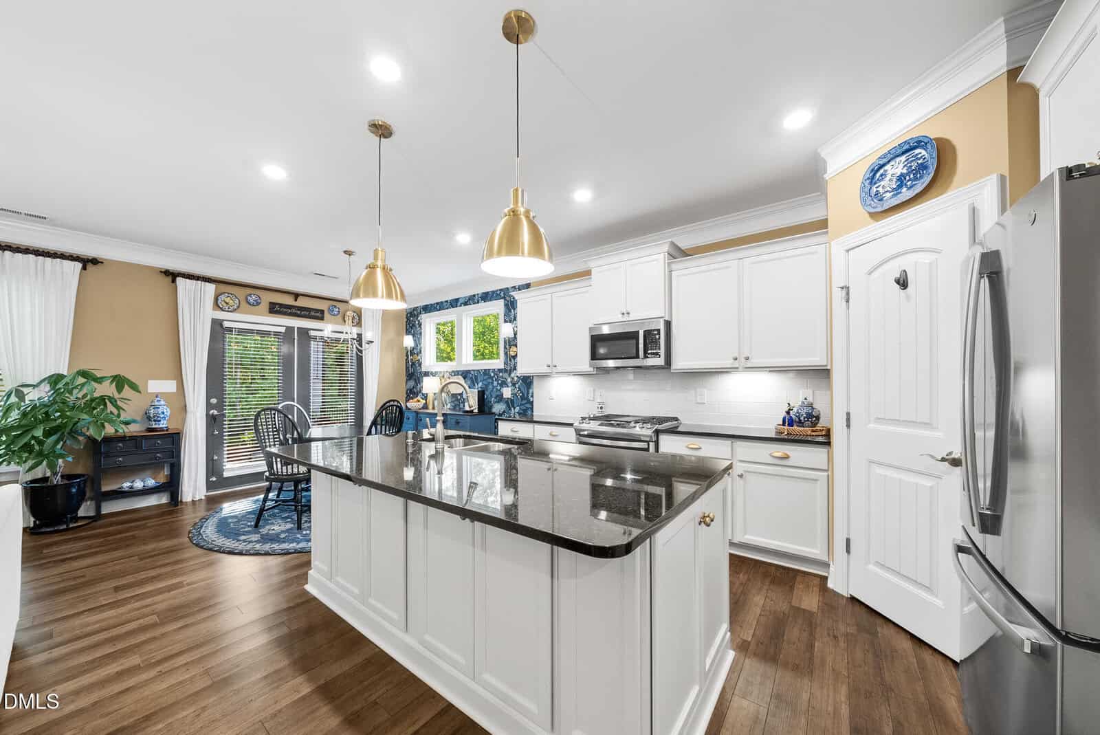 Modern kitchen with white cabinets, stainless steel appliances, a large island with dark countertop, wood flooring, blue and white decor, and dining area with large windows and glass doors to the outside. Two pendant lights hang above the island.