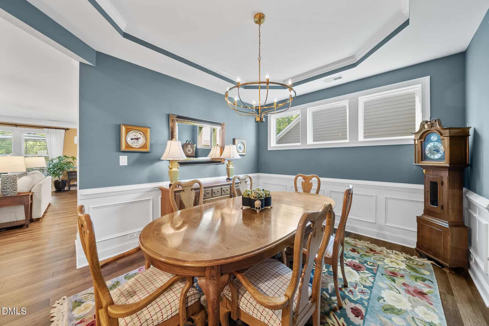 Elegant dining room with blue walls, wooden table and chairs, a floral rug, chandelier, sideboard with lamps, wall art, and a tall grandfather clock. Large windows let in natural light. Living area visible in the background.