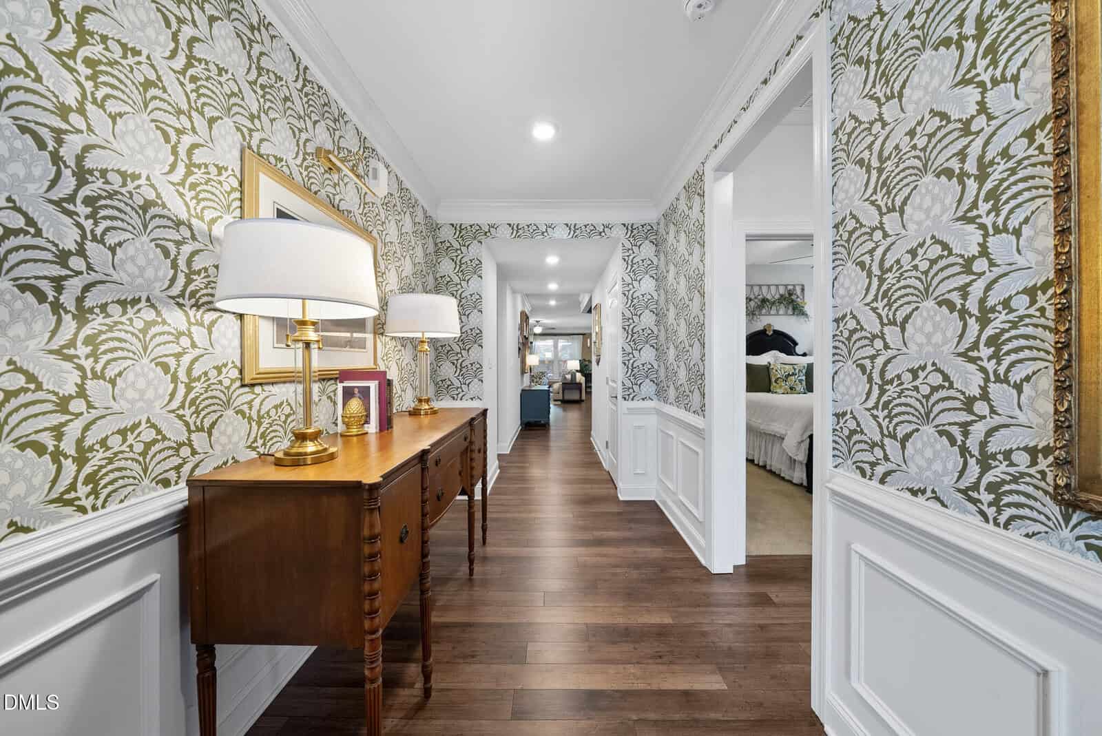 A hallway with floral wallpaper, wooden flooring, and white wainscoting. A wooden console table with lamps and decor stands against the wall; doorways lead to a bedroom and an open living space.