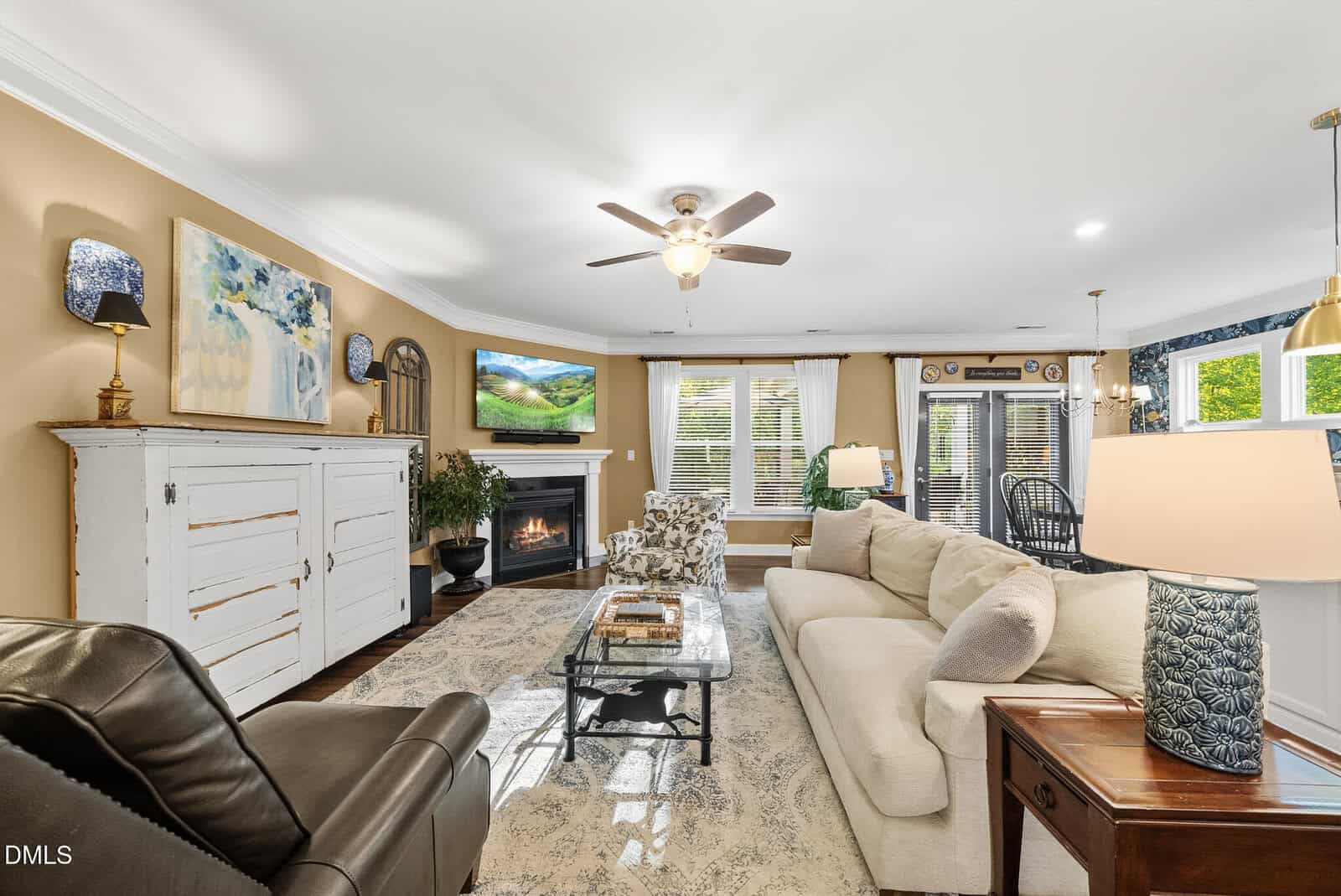 Cozy living room with beige walls, a white couch, glass coffee table, armchair, fireplace with TV above, large rug, and windows with white curtains letting in natural light. Lamps and art decorate the space.