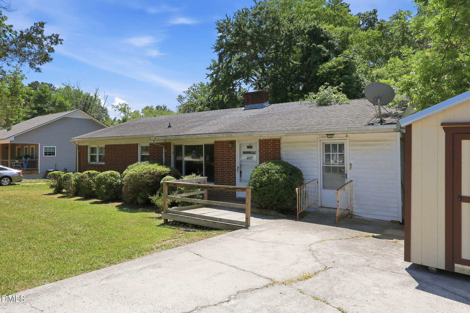 Single-story brick and white siding house with a ramp and steps leading to the front door, bushes along the front, a grassy yard, and a driveway in the foreground. Trees and a neighboring house are in the background.